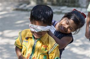 The photo depicts two young children, possibly of Asian descent, engaging in a playful activity. The boy is wearing a bright yellow shirt and has a cloth blindfold tied around his eyes, suggesting they might be playing a game like "blind man's bluff." The girl, with a big smile, appears to be guiding or teasing him, as she holds onto his shoulder from behind. Both children are radiating joy and innocence, and the image conveys a sense of camaraderie and trust. The setting seems to be outdoors in a place with trees and sunlight. This scene is a beautiful representation of childhood, friendship, and the universal language of play that transcends backgrounds and abilities.
