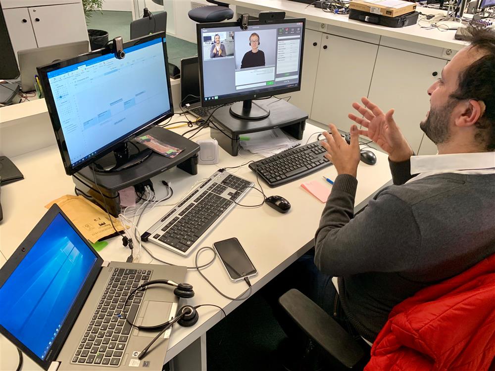 A man sits at a desk with two computer screens in front of him. He is communicating in sign language. On the screen, a sign language interpreter is visible via a video link.  