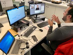 A man sits at a desk with two computer screens in front of him. He is communicating in sign language. On the screen, a sign language interpreter is visible via a video link.  