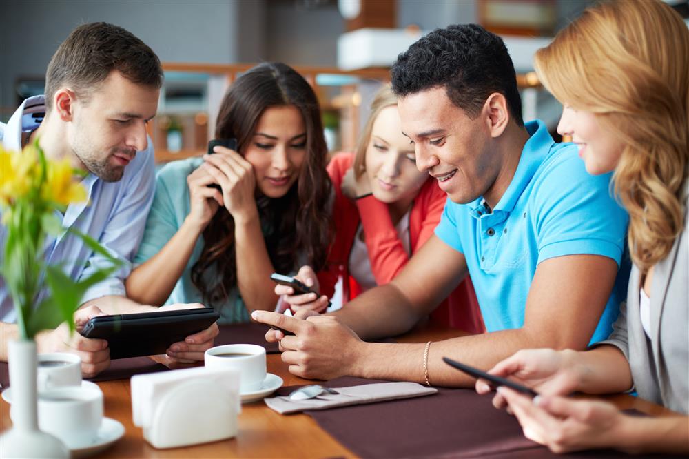 A group of friends sits close together in a cafe and converses via streamer devices, which they are holding in their hands.
