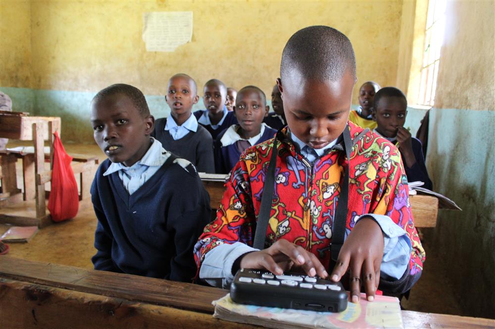 This is an image of a classroom scene with several young African students. In the foreground, a focused student is using a Braille device to read or write, indicating an inclusive educational setting that accommodates visual impairments. The other students in the background appear attentive and engaged. The classroom environment suggests a commitment to providing education and support to all children, regardless of their abilities, promoting themes of equality and assistance.