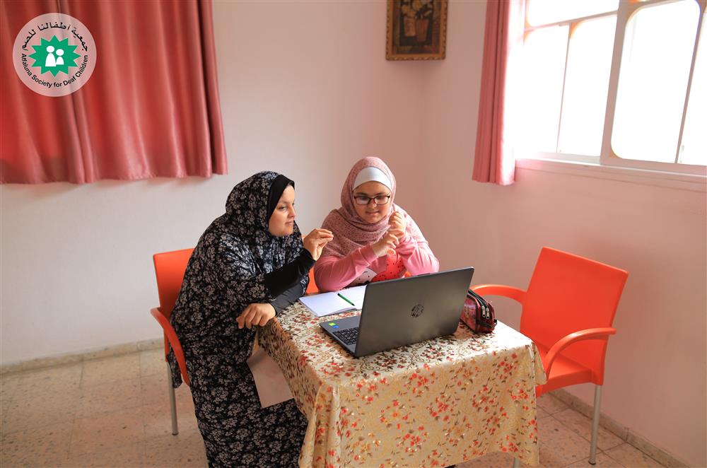 In the image, two women are seated at a table with a floral tablecloth, engaged in what appears to be a collaborative activity. Both women are wearing hijabs, suggesting they may be of a culture where this is a common practice, possibly indicating Middle Eastern or Islamic heritage. The woman on the left is standing and leaning towards the laptop screen, gesturing as if explaining or discussing something with her seated companion. The other woman, seated in front of the laptop, is looking at the screen, with her hands in a position that suggests she may be using sign language, potentially indicating that she is communicating in a way that accommodates a hearing impairment. The setting is a simple room with a window, a pink curtain, and a wall emblem that suggests the environment is dedicated to support or education, possibly a resource center for individuals with hearing disabilities. The atmosphere is one of focus, learning, and mutual assistance.
