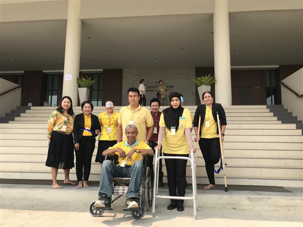 A group picture with a doctor, a volunteer, an engineer, a person with disability and family members standing in front of a white flight of stairs. 