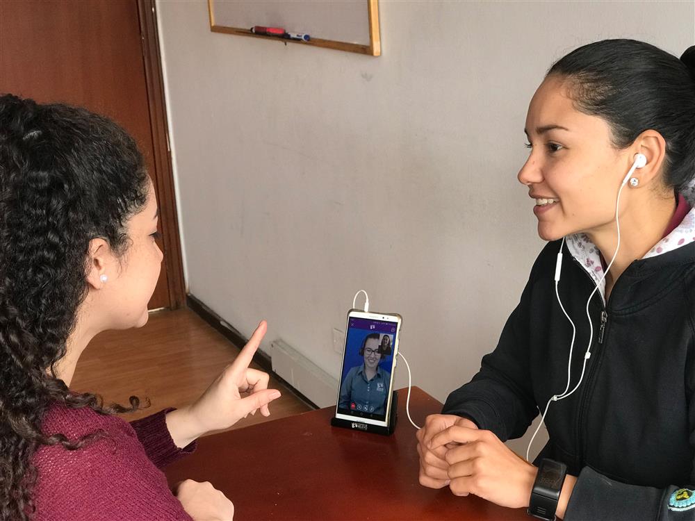 Two young women are engaged in a conversation. The person to the right is connected to a mobile phone. On the screen is a another women wearing headphones. The person to the left makes a gesture