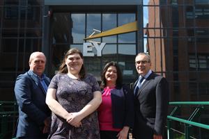 Four people stand outside a building with the Ernst & Young logo. From L-R: man in a grey suit, young woman in a floral dress (Margaret), woman in a navy suit and pink top, man in a black pinstripe suit.