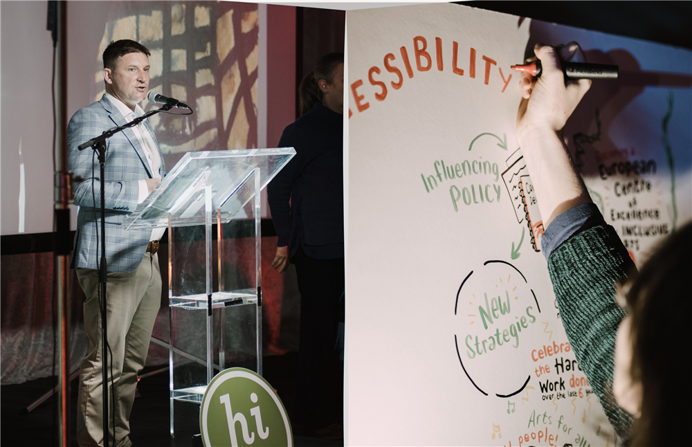 A speaker presents at an event promoting inclusive arts, while another person visually records ideas on a board. Words like “ACCESSIBILITY” and “New Strategies” emphasize collaboration, creativity, and advocacy for equal participation in cultural life.