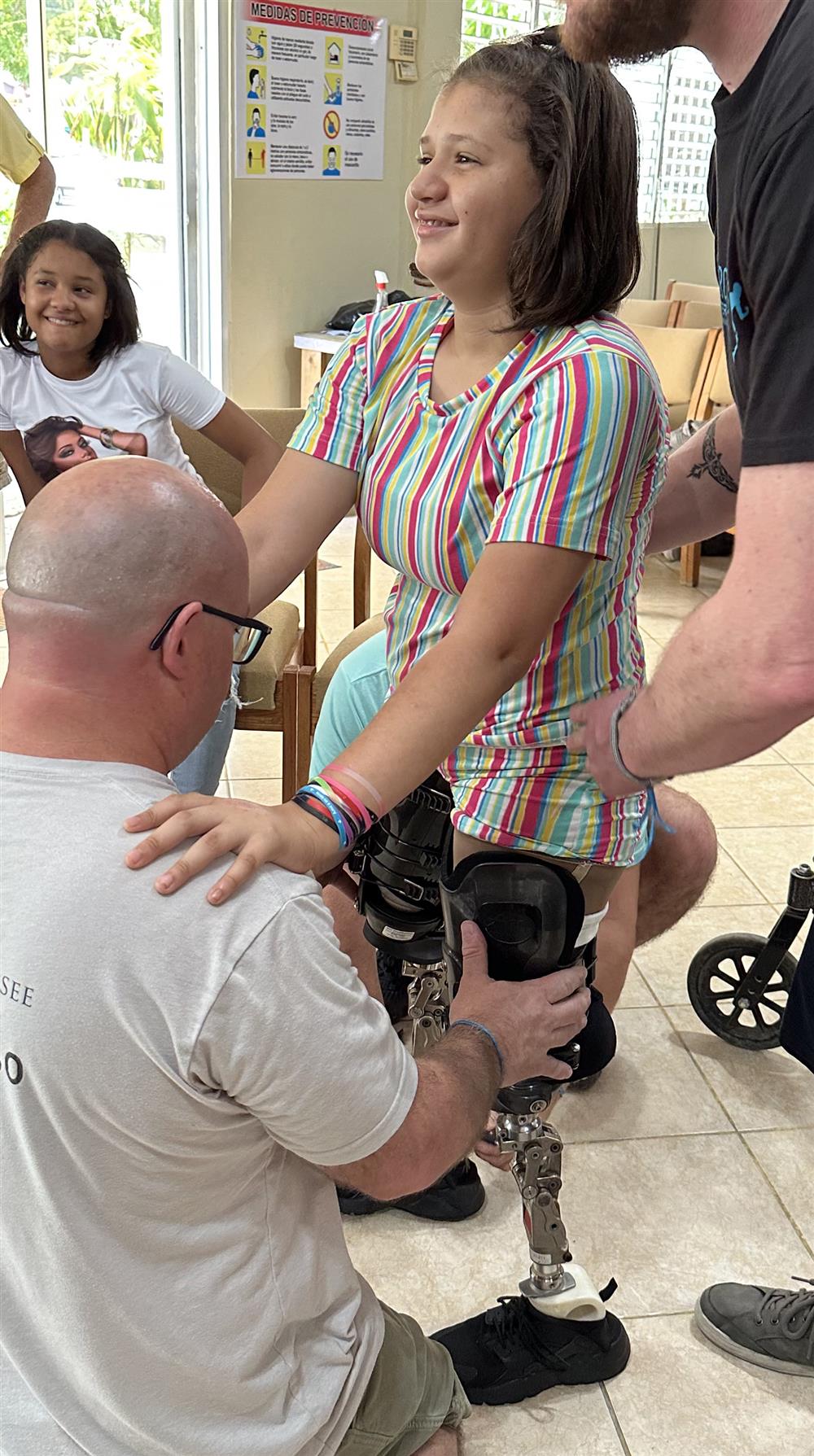 A girl with prosthetic legs smiles brightly as rehabilitation specialists support her in standing. Surrounded by encouragement, her determination and joy embody resilience, inclusion, and the transformative power of assistive technology and compassionate care.