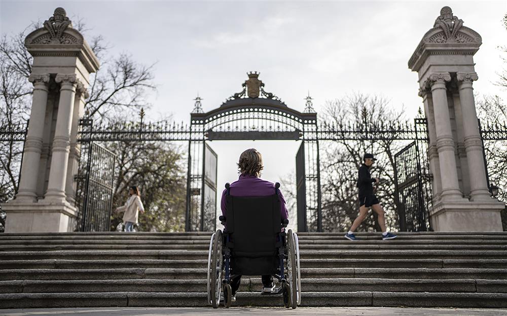 A woman in a wheelchair is facing stairs at the entrance to a park in Madrid. She is turning her back to the audience and it becomes obvious that that stairs are an unsurmountable barrier for her.