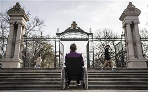 A woman in a wheelchair is facing stairs at the entrance to a park in Madrid. She is turning her back to the audience and it becomes obvious that that stairs are an unsurmountable barrier for her.