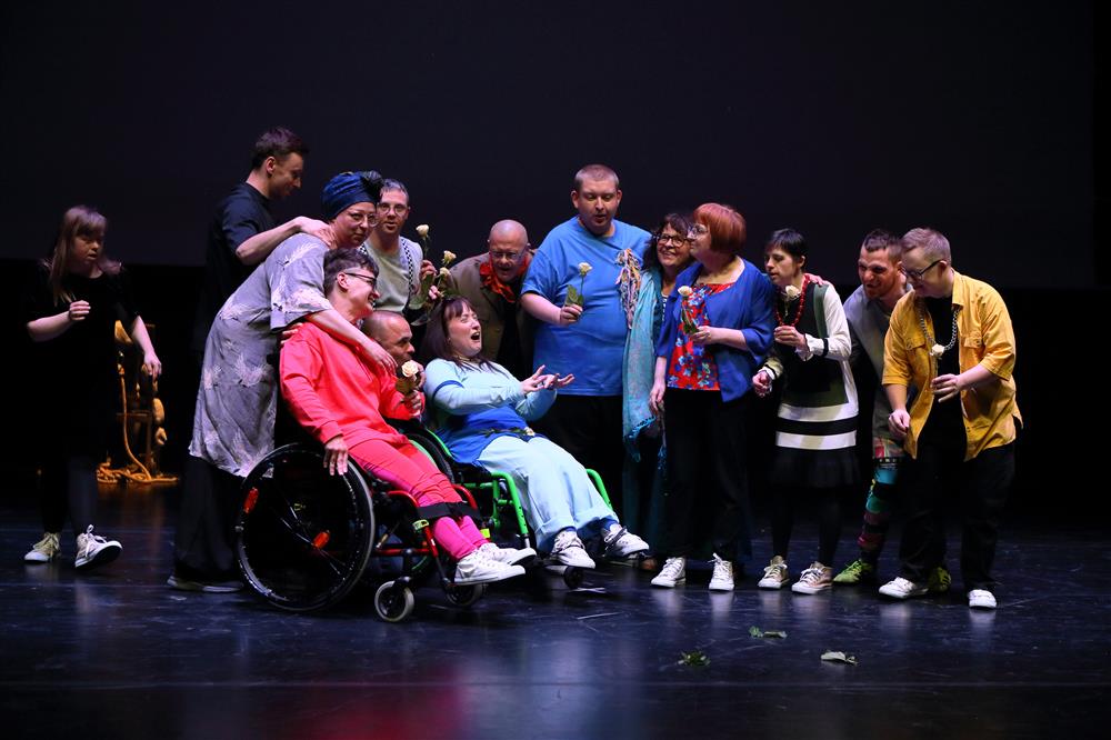 A diverse group of performers, including people with disabilities, gather joyfully on stage holding flowers. Their expressions radiate unity and celebration, embodying how the arts bring people together in creativity, equality, and shared humanity.