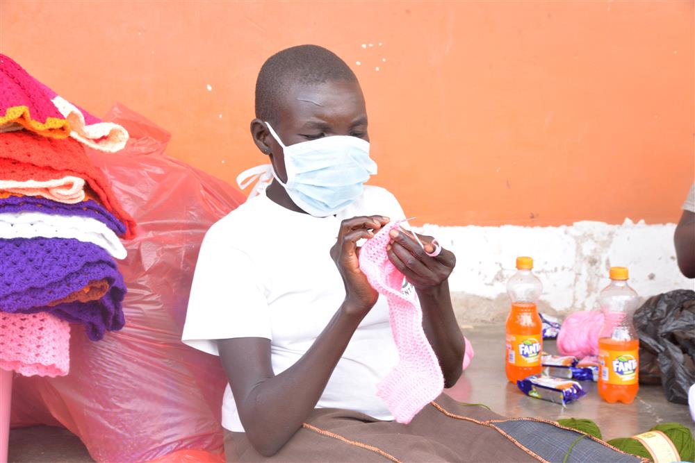 A young woman is sat on the floor leaning against a bag of crocheted goods and with goods piled beside her. She is crocheting using a crochet hook and light pink wool. She is wearing a hygiene mask.