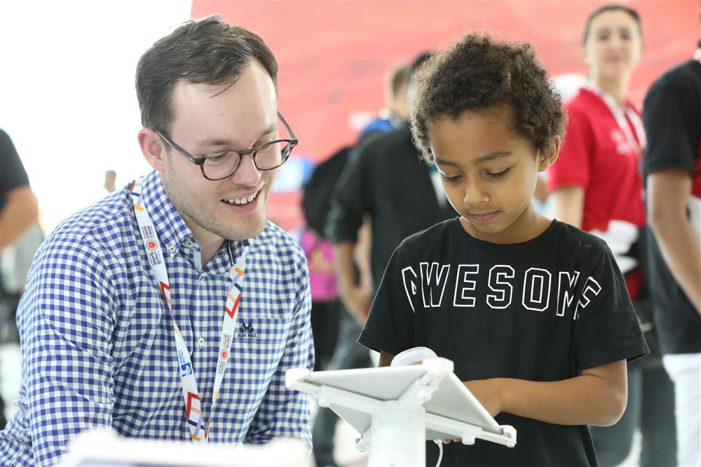 A man sits on a chair next to a boy who is standing. Both are looking at the screen of a tablet in front of them. The boy is touching the screen.