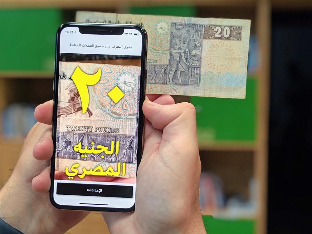 This picture shows someone's hands holding a smartphone on which an app is identifying the banknote held in front of the camera. 