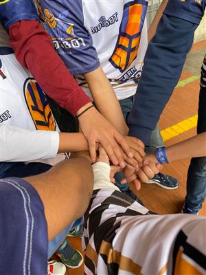 The image shows a group of individuals, likely young students, with their hands joined together in the center of a circle, symbolizing unity and teamwork. They are wearing jerseys with a logo that includes "Team 6301" and "Colombia," suggesting they may be part of a team from Colombia. The variety of skin tones and the cooperative spirit reflect themes of diversity, equality, and solidarity. The act of joining hands is a universal gesture of commitment and support, embodying the values of assistance and justice within a community or team setting.