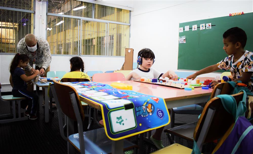 This image depicts a classroom setting with children of various backgrounds engaged in learning activities. An adult, possibly a teacher, is assisting a young girl with her work, emphasizing the importance of support and guidance in education. Another child is wearing headphones, which could suggest an accommodation for a sensory or learning difference, reflecting inclusivity and the adaptation of educational environments to meet diverse needs. The children are interacting with educational toys and materials, illustrating a collaborative and interactive learning space that fosters development and understanding among students. The presence of colorful furniture and a chalkboard suggests an environment designed for young learners.