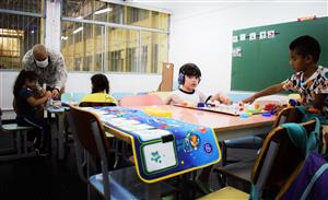 This image depicts a classroom setting with children of various backgrounds engaged in learning activities. An adult, possibly a teacher, is assisting a young girl with her work, emphasizing the importance of support and guidance in education. Another child is wearing headphones, which could suggest an accommodation for a sensory or learning difference, reflecting inclusivity and the adaptation of educational environments to meet diverse needs. The children are interacting with educational toys and materials, illustrating a collaborative and interactive learning space that fosters development and understanding among students. The presence of colorful furniture and a chalkboard suggests an environment designed for young learners.