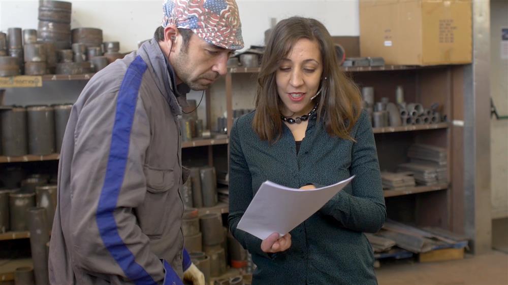 A man and a woman stand next to each other in a workshop with shelves of metal pipes behind them. The woman is showing the man something on a piece of paper. 