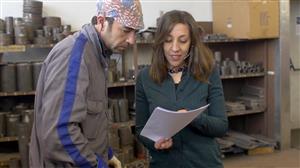 A man and a woman stand next to each other in a workshop with shelves of metal pipes behind them. The woman is showing the man something on a piece of paper. 