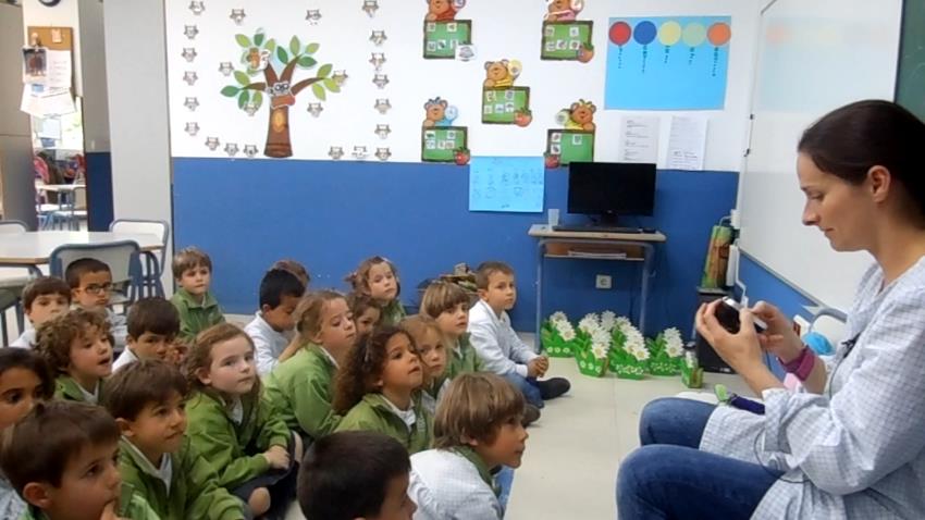 Young children are sitting on the floor and looking up ito their teacher, who holds a device.