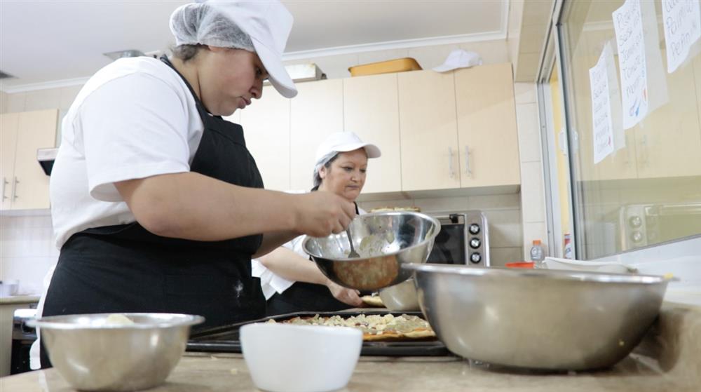 Two women are in a kitchen in a pizzeria. One woman holds a spoon and is spooning ingredients from a bowl onto a pizza base while the other woman observes her.