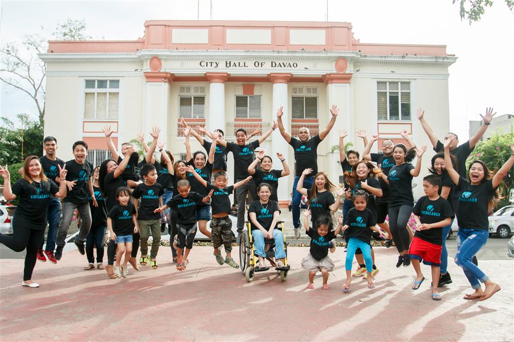 There are 16 people wearing a Virtualahan shirt jumping with two people who are in their wheelchairs in front raising their hands enthusiastically.