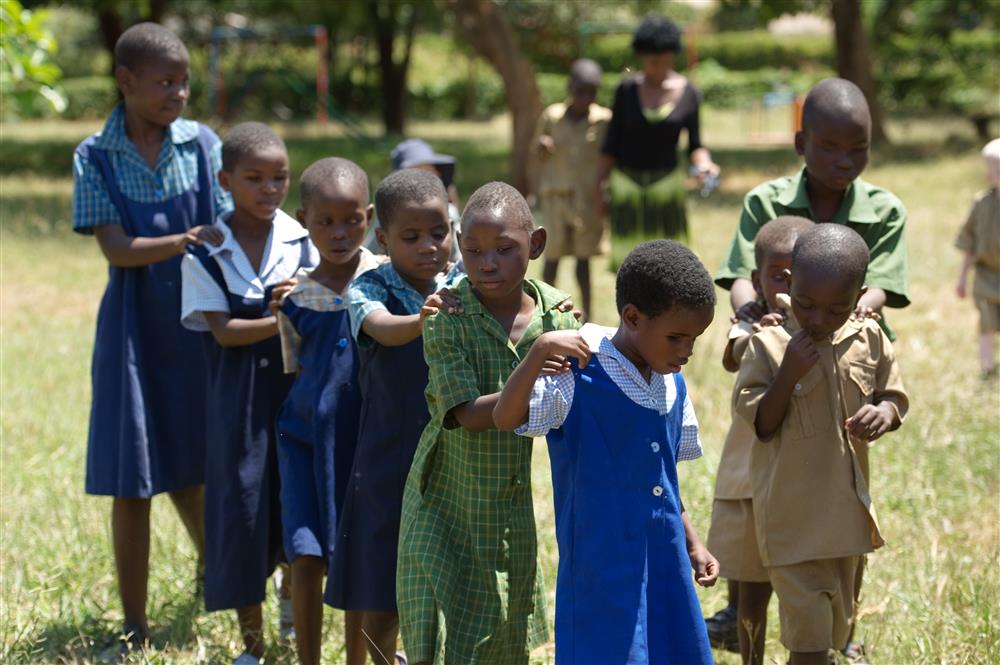Children in school uniforms walk outside, grapping each other by the shoulders to be leaded back to the classroom.