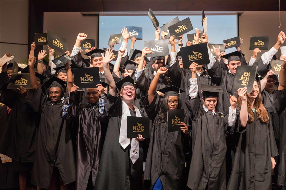 A group of graduates in ropes are waving their hands and holding their graduation certificate up.