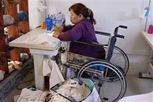 A woman in a wheelchair sits at a sewing machine sewing a canvas tote bag. A large stack of finished totes is on the floor nearby.