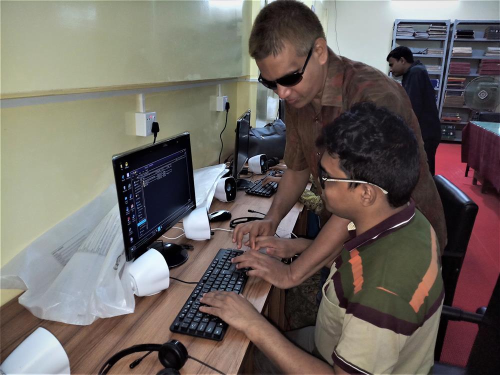 Set in a classroom of an accessbile e-learning center, a blind teacher wearing sunglases explains how to use the keyboard  to a young blind boy  in order to access an e-learning program on his screen.