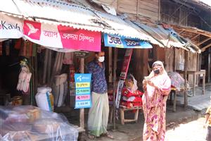 In a rural marketplace, a woman in traditional South Asian dress counts money outside a small shop, while another person stands nearby. The scene reflects economic empowerment, community connection, and access to digital and financial inclusion in local settings.