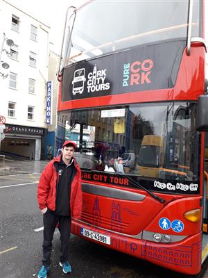 A young man in a black tracksuit with a red cap and rainproof jacket stands in front of a tour bus with Cork City Tours written on it. He is looking at the camera and smiling.