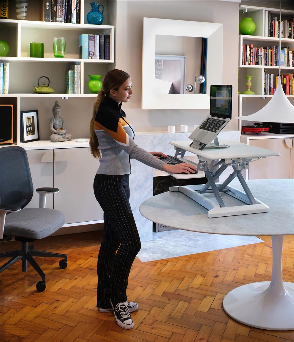 A young woman is standing at a desk looking at a computer screen. The desk is height adjustable. In the background are an office chair and a modern design shelf.