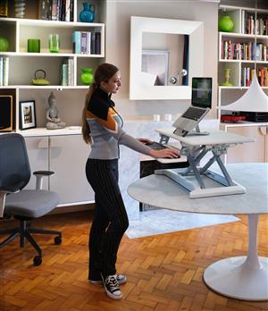 A young woman is standing at a desk looking at a computer screen. The desk is height adjustable. In the background are an office chair and a modern design shelf.