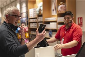 A grey haired man holds up a mobile phone and makes a gesture with his hands. In the background is a shop assistant watching the scene.