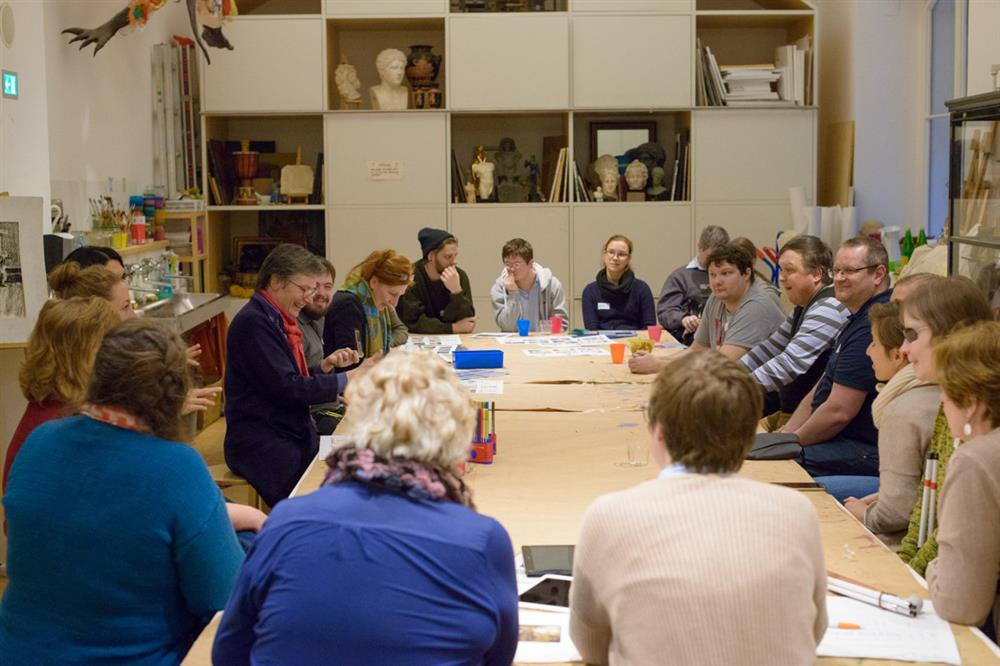 Project participants are sitting around a table in front a shelf, which holds art works.