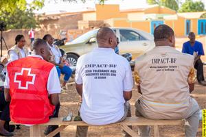 Humanitarian workers, including members of the Red Cross and IMPACT-BF, sit together outdoors. Their shirts display messages advocating zero tolerance for gender-based violence and inclusive humanitarian action—promoting dignity, equality, and justice for all.