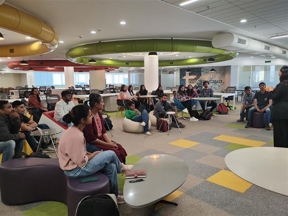 The photo depicts a diverse group of individuals attentively gathered in a modern indoor setting, which appears to be a workspace or educational environment. They are seated on various types of furniture, including bean bags and chairs, arranged in a semi-circle around a standing speaker, suggesting an informal meeting or learning session. The room is well-lit and features colorful decor, with circular light fixtures and vibrant carpet tiles. The atmosphere seems inclusive and collaborative, highlighting themes of community and shared learning.
