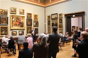 A tour guide in front of many Renaissance paintings gives a tour to visiters, who are sitting or standing in a big exibition room of the Staatlichen Museum Schwerin.