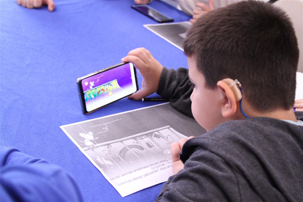 A boy wearing a hearing aid is looking at the mobile screen with a paper that has a similar image printed in a paper. 