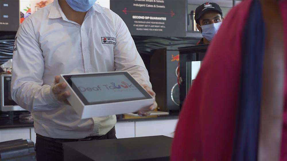 A man stands behind the counter at a coffee shop and holds a tablet displaying the DeafTawk app to a woman on the other side of the counter.