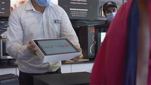 A man stands behind the counter at a coffee shop and holds a tablet displaying the DeafTawk app to a woman on the other side of the counter.
