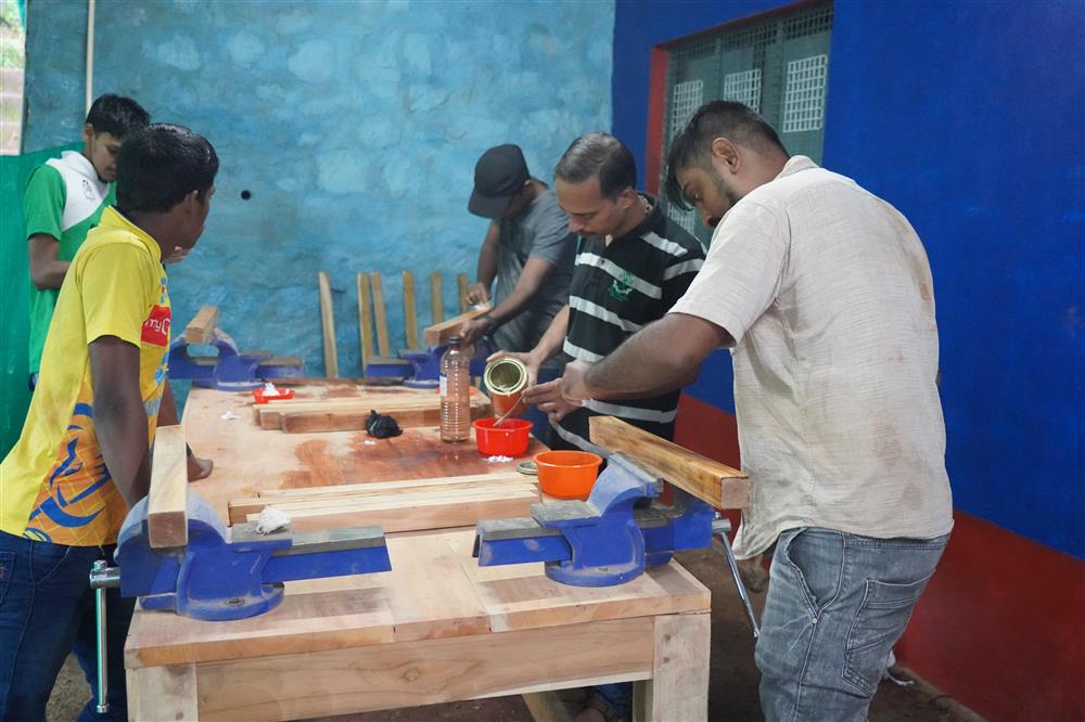 The photo shows a group of individuals engaged in a woodworking activity. They are working together at a sturdy workbench equipped with vices to hold the wood in place. One person is pouring a liquid, possibly a finish or adhesive, into a container, while others are observing or waiting their turn. The setting suggests a workshop environment, with a focus on collaboration and skill-sharing. The image conveys themes of cooperation, learning, and community engagement. The individuals' attire is casual, indicating a relaxed and informal atmosphere conducive to hands-on learning and teamwork.