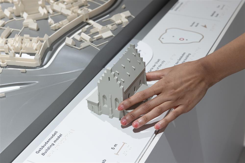 A hand gently explores a tactile architectural model, part of an accessible exhibit designed for inclusive learning. The display includes braille text, symbolizing equal access to culture and knowledge for people with visual impairments.