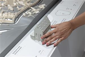 A hand gently explores a tactile architectural model, part of an accessible exhibit designed for inclusive learning. The display includes braille text, symbolizing equal access to culture and knowledge for people with visual impairments.