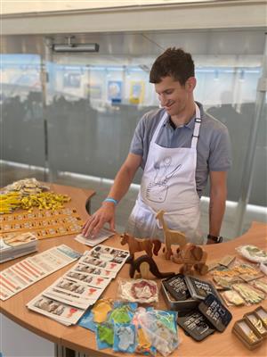 A man wearing apron touching cards on the table. Different figurines, pins, candies and stickers are arranged on the table to be sold.