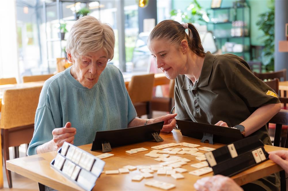 An elderly woman and a younger companion enjoy a game together, deeply focused on the board. The scene reflects connection across generations, demonstrating patience, empathy, and shared joy in a community setting.