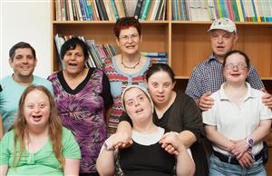 Six individuals of different races who appears to have intellectual disabilities standing in front of  a large bookshelf half-filled with books. One woman in the group has her arms around the shoulder of one of the two women who appears to also have intellectual disability sitting in front of the group.