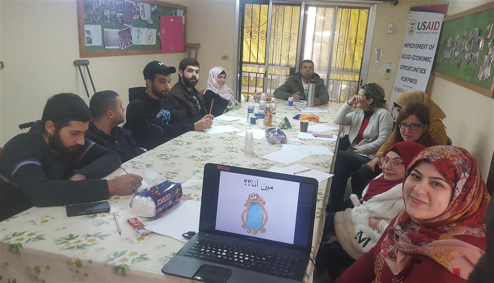 There is a long rectangular table with five young people sitting on each side and one person at the head. A laptop and training materials are on the table. Most people are  smiling.