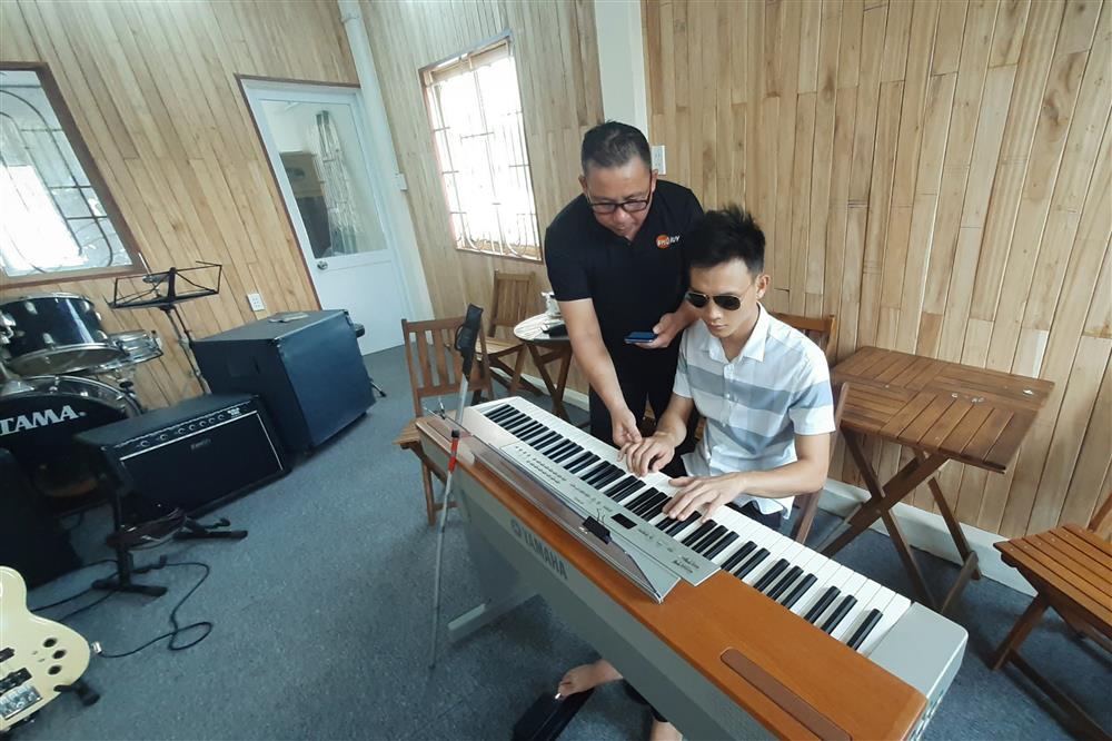 A blind students plays an electronic piano. His teacher stands next to him and uses a mobile phone with the SM Music app. There is music equipment visible in the background of the room.