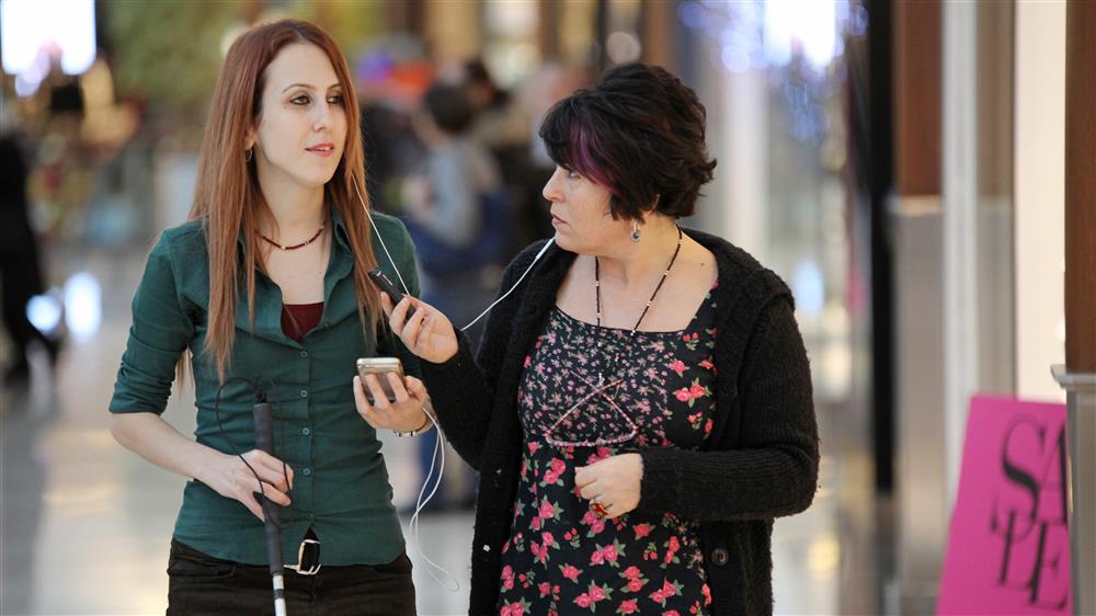 Two women are in a shopping mall. They are sharing headphones and one of them is holding a mobile phone. The person to the left holds a white cane. 
